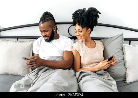Joyful positive African American couple, happy spouses, wife and husband sit in a cozy bed in the bedroom, use their smartphones, browsing social networks, news, messaging with friends, smile Stock Photo