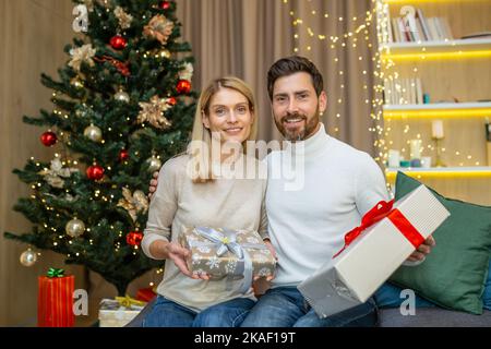 Portrait of a happy young couple celebrating Christmas at home. They are sitting on the sofa near the Christmas tree with gifts in their hands, looking at the camera, smiling. Stock Photo