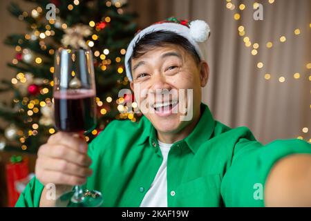 Happy young handsome Asian man in red Santa Claus hat and green shirt taking selfie at home near Christmas tree, holding glass of wine, smiling. Stock Photo