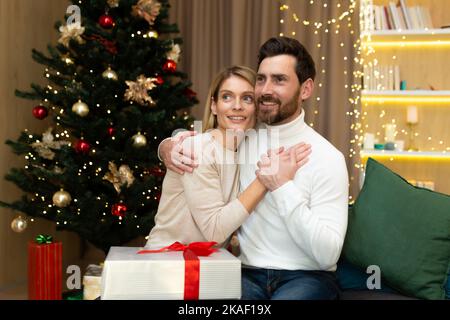 Portrait of a happy young couple, family. Young man and woman celebrating Christmas holidays at home. They sit on the sofa near the Christmas tree with gifts, hug, rejoice, look away. Stock Photo