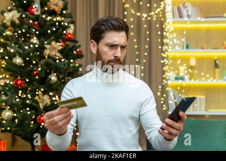 Confused and embarrassed man looks at the phone, holds a credit card in his hands. Sitting on the sofa near the Christmas tree, unable to make festive online purchases, he doesn't have enough money. Stock Photo