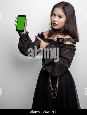 A Filipina in a vampire costume pointing at a phone with a green screen on white background Stock Photo