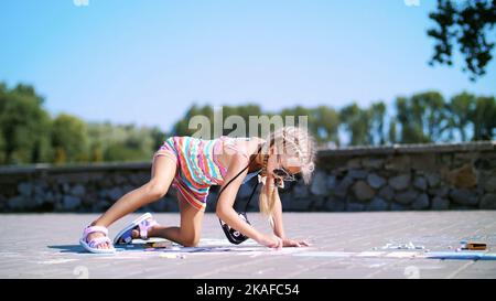 a girl in sunglasses, draws drawings with colored crayons on the asphalt, street tiles. A hot summer day. High quality photo Stock Photo