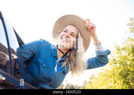 Young woman wearing blue denim shirt, holding beige floppy hat, sitting in black car leaning out of window in sunny day. Stock Photo