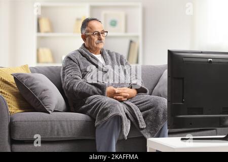 Mature man wearing a bathrobe, sitting on a gray sofa and watchin tv at home Stock Photo