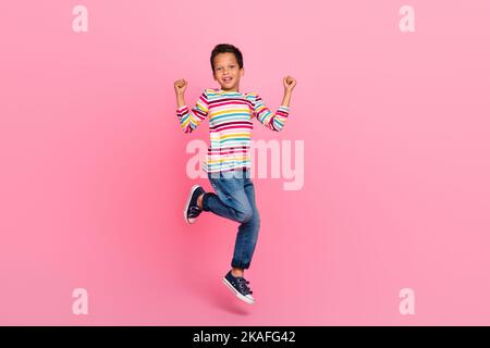 Full length photo of cheerful optimistic adorable boy dressed striped shirt jeans jumping hold fists up isolated on pink color background Stock Photo