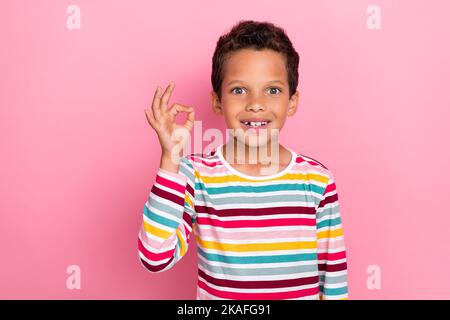 Portrait of adorable charming boy with wavy hairstyle wear striped long sleeve showing okey sign symbol isolated on pink color background Stock Photo