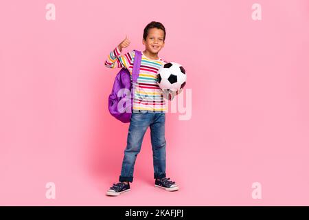 Full size photo of charming friendly cheerful boy striped shirt bag pack holding soccer ball show like isolated on pink color background Stock Photo