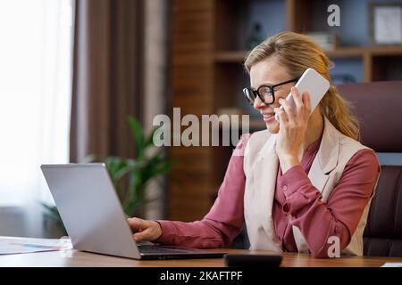 Portrait of smiling businesswoman talking on the phone using laptop in office interior. Female office worker searching new clients, negotiates with Stock Photo