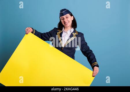 Smiling stewardess holding yellow empty advertising banner with copy space. Happy flight attendant, standing with blank placard in hands. Air hostess in aviation uniform with promotion mockup Stock Photo