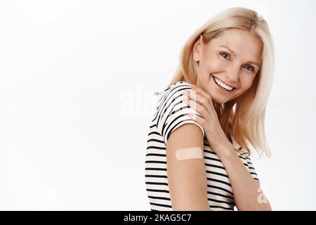 Happy and healthy mature woman, 50 years old, has band aid on shoulder, smiling and laughing, standing in tshirt over white background. Stock Photo