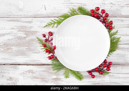 Christmas or winter table setting mock up. Blank white plate with frame of evergreen branches and red berries. Above view on a white wood background. Stock Photo