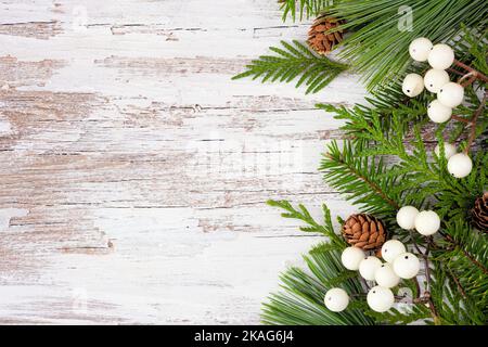 Winter side border with evergreen branches, white berries and pine cones. Overhead view on a rustic white wood background with copy space. Stock Photo
