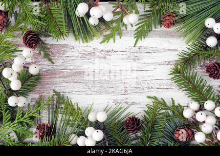 Winter frame with evergreen branches, white berries and pine cones. Top down view on a rustic white wood banner background with copy space. Stock Photo