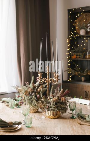 Christmas and New Year's decor of the dining table: candles in a classic candlestick, glasses, compositions of fir branches in golden vases Stock Photo