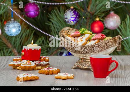 Silver vase with gingerbread cookies, a burning candle, a cup of coffee and christmas fir tree with toy balls and garlands on the background. Stock Photo