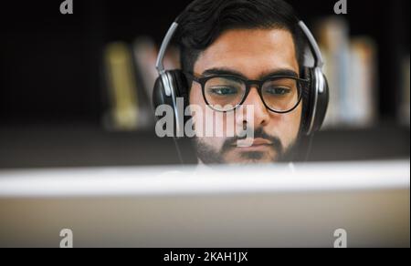 The music will get me trough this. Closeup shot of a focused young man sitting and working on a computer in a library while listening to music. Stock Photo