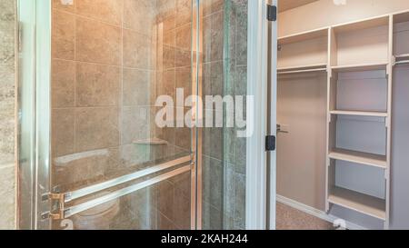 Panorama Walk-in closet with shelving units near the shower stall. There is a shower stall with gray tiles surround and glass door with aluminum frame Stock Photo