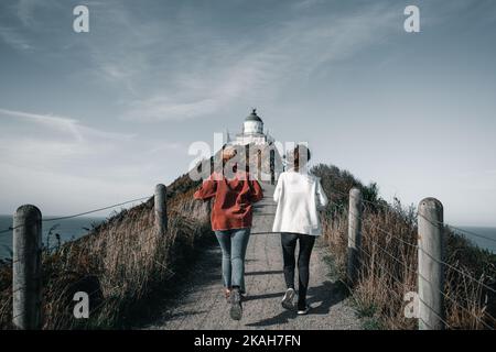 two caucasian girls in red jacket hat and white fleece sweater and sneakers are running towards the impressive lighthouse along the dirt road, nugget Stock Photo