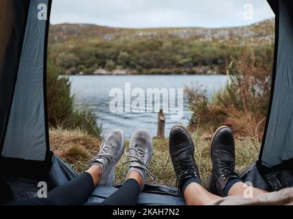 Shoes, camping and tent with a couple by a lake out in nature while hiking for adventure or travel together. Water, forest and freedom with a man and Stock Photo