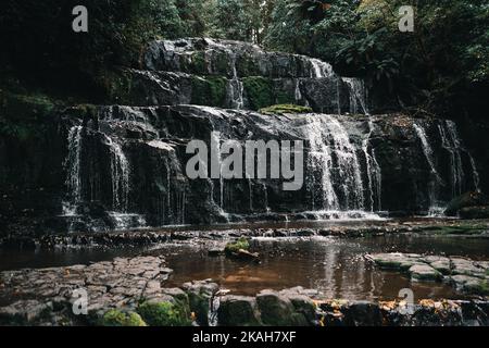 beautiful waterfalls falling down the big rocks among the greenery of the forest trees near the small lake with a lot of stones in the middle of the Stock Photo