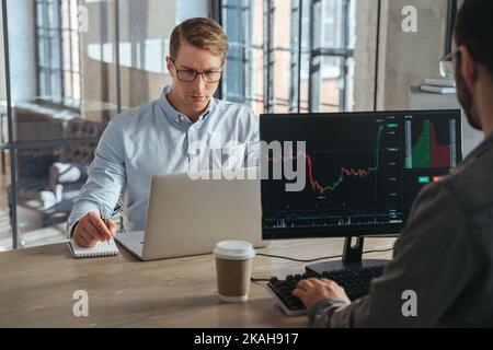 Two stock traders co-working at office sitting at table in front of computers opposite each other, working online, analyzing stock exchange market tendency, weighing risks, selling or buying crypto Stock Photo