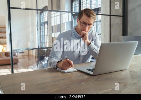 Handsome businessman in shirt and glasses sitting at office table in front of laptop, making notes in notepad, planning meetings with investors.Crypto trader analysis stock market. Copy space Stock Photo