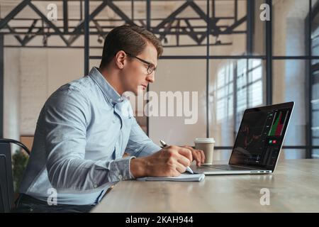 Busy male trader in glasses working online on laptop at his office, financial crypto expert analyzing graph chart looking at screen of monitor with diagram, writing in notepad. Cryptocurrency Stock Photo