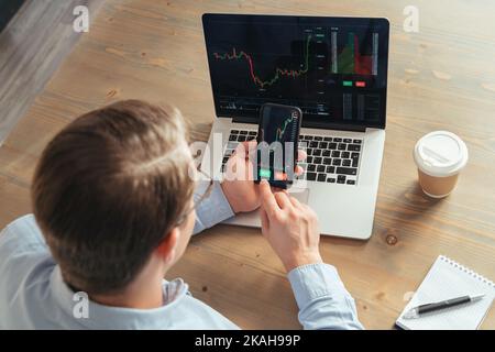 Over shoulder top view of business man trader investor using smartphone cryptocurrency app financial market analysis, trading data index chart with buy and sell buttons Stock Photo
