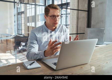Handsome financial coach having online lesson with his student teaching basics of investments, trading strategy and cryptocurrency, sitting at his office in front of laptop, talking and gesticulating Stock Photo