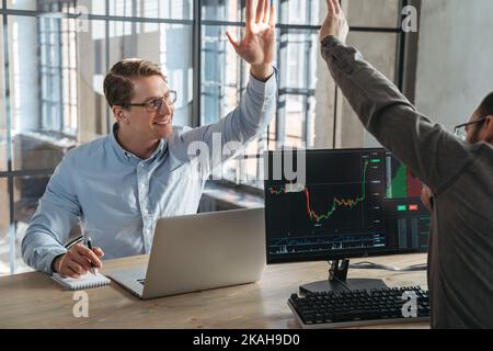 Two partners trade investors giving high five to each other after fulfilling good deal, buying crypto currency at stock market at low price, sitting opposite each other in front of computers Stock Photo