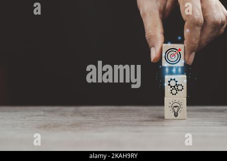 Goals Setting. Female Hand Putting Wooden Blocks With Picture Of Target, Working Gearwheels And Lightbulb Over dark  Background. Goal Achievement Conc Stock Photo