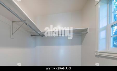 Panorama Small empty walk in closet with white interior and window. There is a carpeted flooring and shelves on the walls with metal brakets and rods Stock Photo