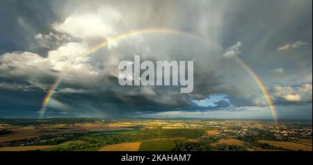 A rainbow over a farmland, the remnants of a storm, a dramatic sky, ripening cornfields, a town and hills in the background. Aerial panoramic view. Stock Photo