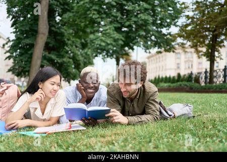 group of students reading a textbook lying on the grass . Stock Photo