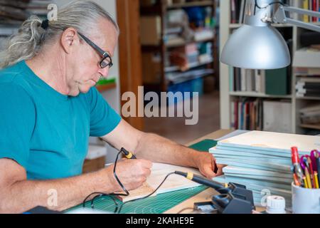 Senior man working with the pyrographer burning the wood making artwork craft. Stock Photo