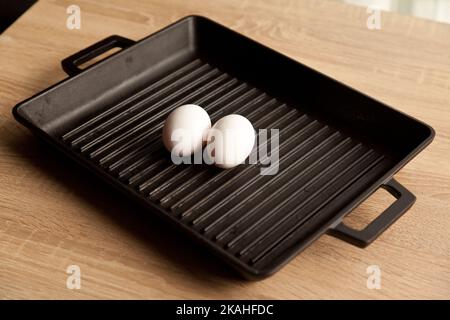 Black cast heavy iron grill pan in two organic white eggs; on modern oak kitchen countertop, modern kitchen equipment Stock Photo