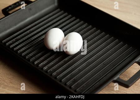 Black cast heavy iron grill pan in two organic white eggs; on modern oak kitchen countertop, modern kitchen equipment Stock Photo