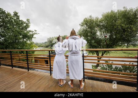 Back of couple in white bath robe and sauna hat stand in wooden terrace of modern house. Stock Photo