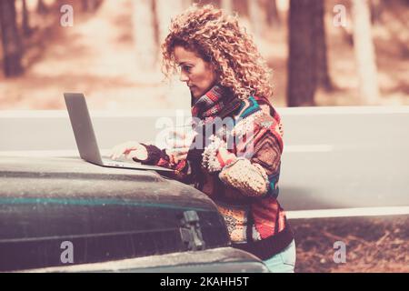 Autumn color and side view of one woman using laptop computer on the car outside with woods and nature in background. Concept of travel people and roa Stock Photo