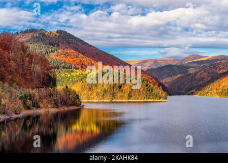 Scenic view of autumn colored forest reflecting to alpine lake in Transylvanian Alps during warm October light against dramatic sky Stock Photo