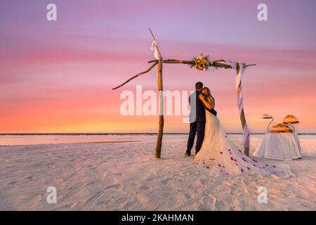 Bride and groom, newlyweds, honeymoon on beach sunset sun romance under wedding arch with cake. Lovers or newlywed married young couple by sea sand Stock Photo