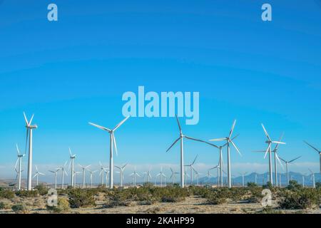 Windmills with mountains and clear blue sky on a sunny day landscape. Windpumps with three blades on a sunlit field generating alternative energy. Stock Photo
