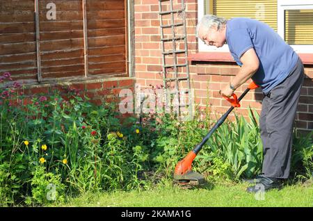 Senior man happy strimming his garden. Using strimmer to cut down grass or weeds. Stock Photo