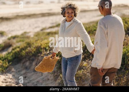 Happy senior woman holding her husband by the hand while going to the beach for a picnic. Cheerful elderly couple taking a seaside holiday after retir Stock Photo