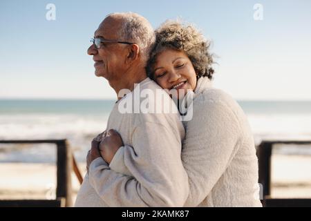 Affectionate senior woman smiling happily while embracing her husband by the ocean. Romantic elderly couple enjoying spending some quality time togeth Stock Photo