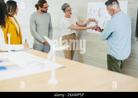 Mature design professional explaining some drawings during a meeting with her colleagues. Innovative businesspeople sharing creative ideas while worki Stock Photo