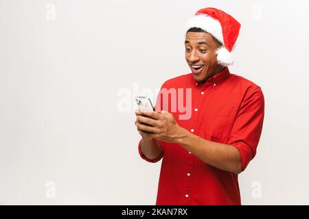 Close-up portrait of hilarious african-american man in santa hat using smartphone isolated on white, young guy sending New Year congratulations, texting online Stock Photo