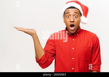 Shocked African-american guy in santa hat and red shirt points with hand aside isolated on white background, astonished guy looks at the camera with wide opened eyes and mouth Stock Photo