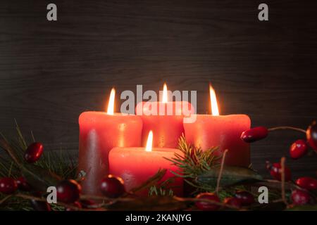 Four red advent candles lit on an advent wreath in a dark room with evergreen boughs and red berries with copy space Stock Photo